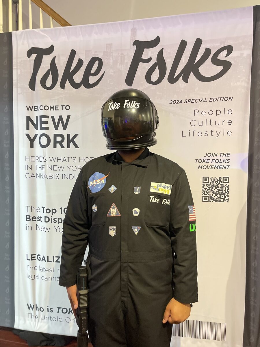 A guest in a flight suit and helmet on the Toke Folks cover backdrop